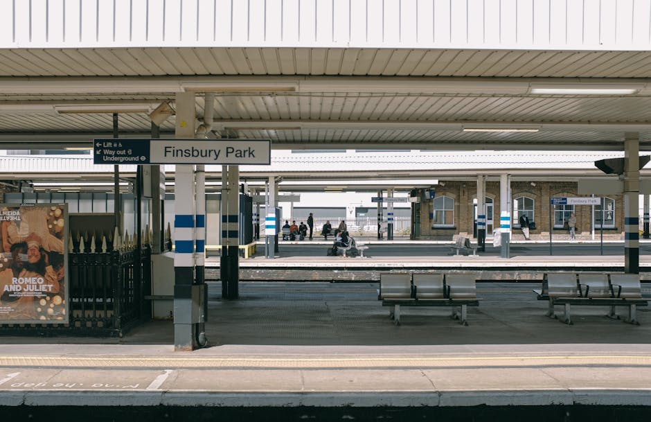 The image shows Finsbury Park railway station platform under a large, cream-colored roof supported by multiple white pillars. There are several wooden benches arranged along the platform, with a few passengers sitting and waiting. In the foreground, a black metal fence surrounds a poster advertising Romeo and Juliet, with a small section of the platform's edge visible, including tactile paving. Further in the background, additional platforms and passengers are visible, with some individuals walking or standing near the edge of the tracks. The station environment appears clean and organized, with signage indicating lift and underground directions visible on the pillars. This scene depicts a typical train station setting, capturing the calm moment before departures, relevant for illustrating home relocation or moving logistics involving train travel, as handled by companies like Man with Van Manor Park.