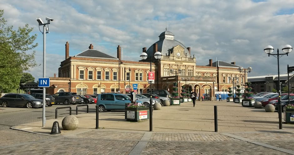A large, historic railway station building made of red brick and stone, featuring tall arched windows and a decorative central clock tower. The foreground shows a paved area with parked cars, including hatchbacks and sedans, arranged along the perimeter of the station. Several black metal bollards and planter boxes with colorful flowers are positioned around the area, with some pedestrians walking nearby. A blue 'IN' traffic sign is visible on a pole to the left. To the right, a decorative lamppost with multiple spherical lights stands next to a group of people. The scene is illuminated by natural daylight, with a partly cloudy sky overhead. The environment appears calm and organized, indicating a public transport hub or busy town centre, which may be relevant for local house removals or furniture transport services in the area, as provided by Man with Van Manor Park.