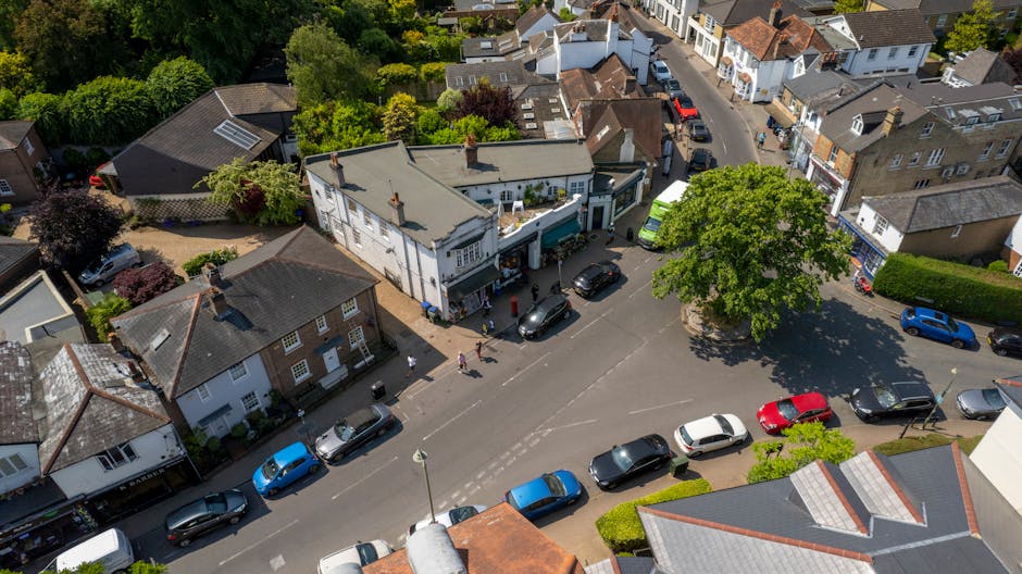 An aerial view of a street scene in Manor Park showing a house with a white facade and a grey roof, positioned on the corner of the street, surrounded by several parked cars including a blue, red, bla
