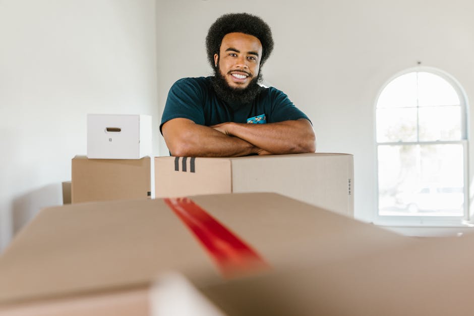 A man wearing a blue jumpsuit and a headband stands inside a room with a textured blue-grey wall and wooden flooring, surrounded by several packed cardboard boxes of various sizes stacked on the floor