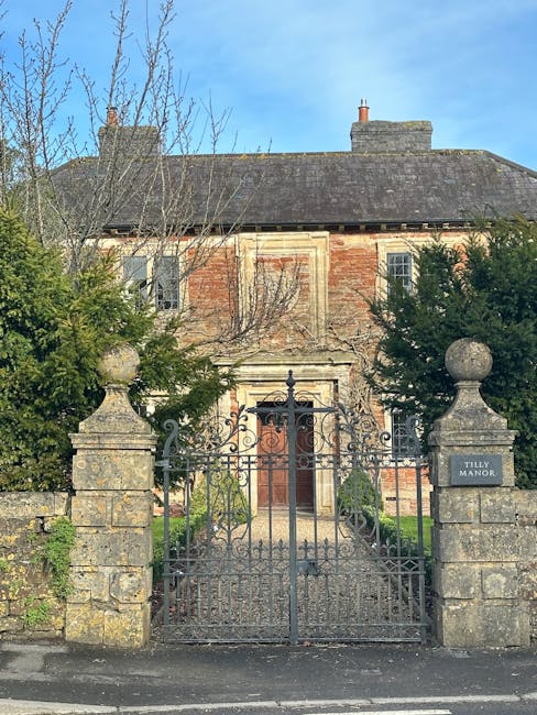 Photograph of a traditional two-storey brick house with a grey slate roof, situated behind a decorative metal gate flanked by stone pillars displaying rounded stone finials. The house features a centr