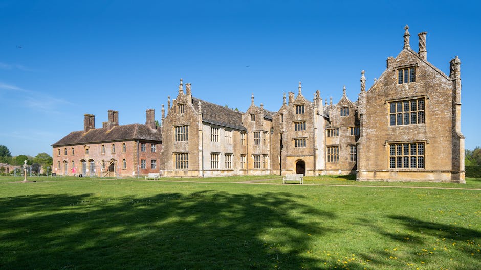 Photograph of a traditional two-storey brick house with a grey slate roof, situated behind a decorative metal gate flanked by stone pillars displaying rounded stone finials. The house features a centr