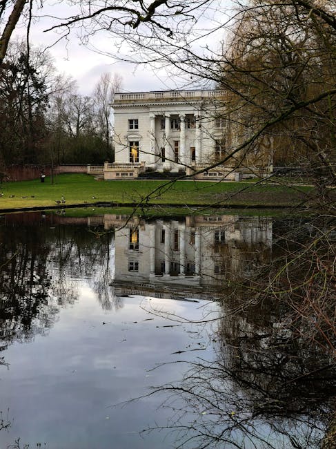 A white, neoclassical-style mansion with tall columns and decorative balustrades on the upper balcony, situated behind a small lake or pond with calm, reflective water. Surrounding the property are leafless trees and a well-maintained grassy lawn. The reflection of the mansion is visible in the water, and the scene is captured on an overcast day with diffused lighting. This image depicts a residential property set in a landscaped environment, relevant to house removals and relocation services provided by Man with Van Manor Park, highlighting the proximity of outdoor spaces and the importance of careful planning during home furniture transport and moving logistics.