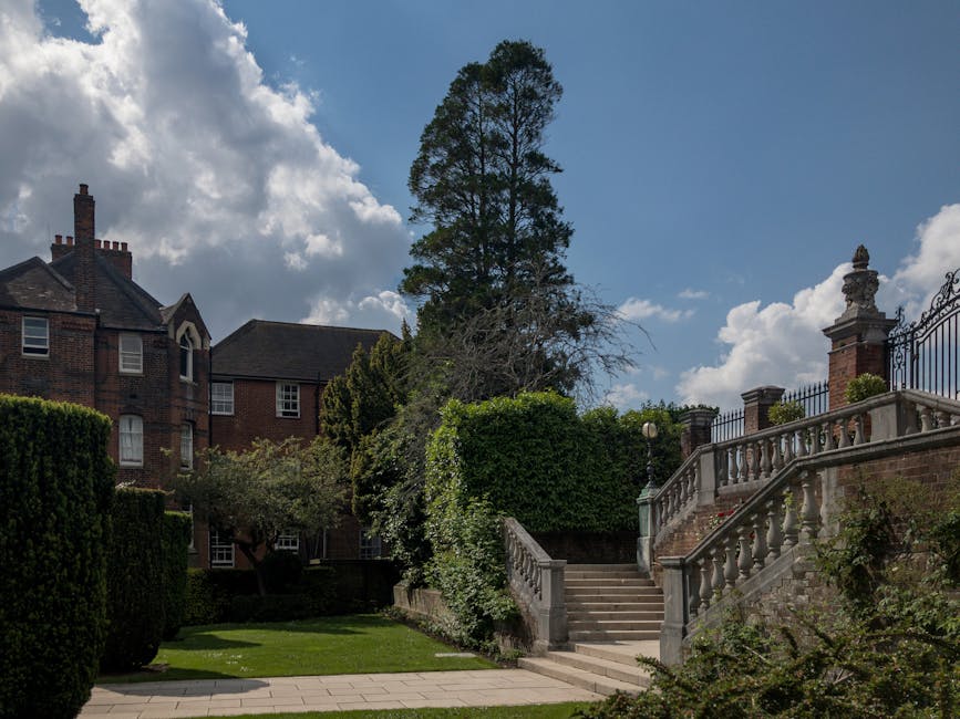 A residential property exterior scene showing a stone staircase with a decorative balustrade leading from a paved pathway up to an ornate brick and stone entrance gate. To the left of the staircase, there are well-maintained, trimmed bushes and a lush green lawn. A tall, mature tree with sparse foliage towers above, set against a partly cloudy sky. Behind the bushes and trees, parts of a large brick building with multiple windows and chimney stacks are visible. The scene captures the outdoor environment used during house removals or home relocation activities, with no moving equipment or vehicles in sight, focusing on the setting for transportation and packing procedures.