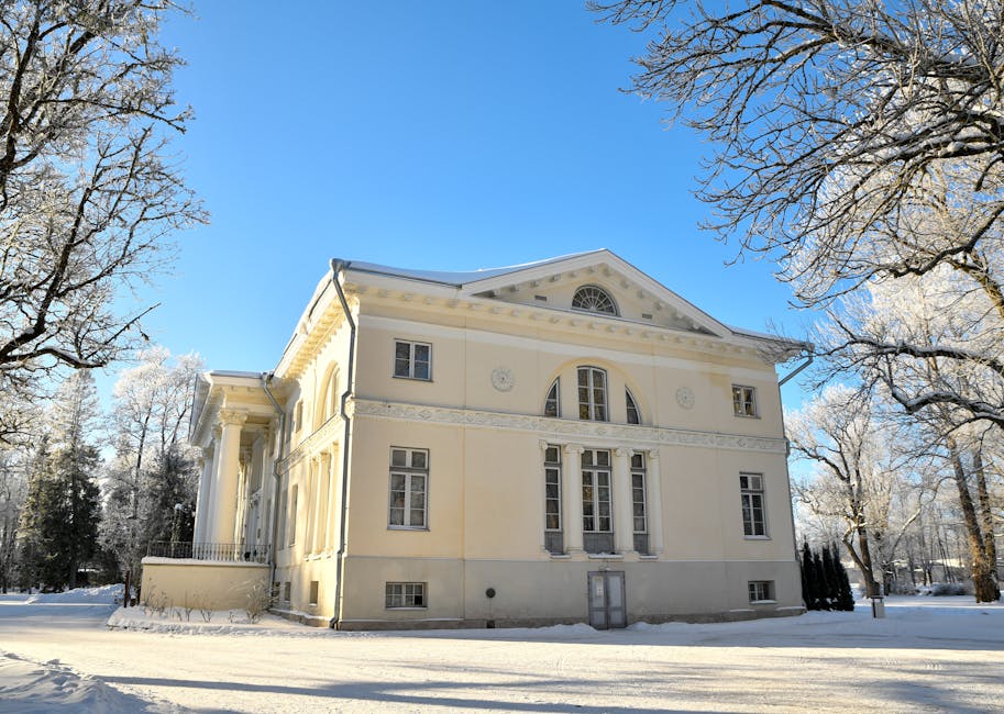 A white, neoclassical-style mansion with tall columns and decorative balustrades on the upper balcony, situated behind a small lake or pond with calm, reflective water. Surrounding the property are leafless trees and a well-maintained grassy lawn. The reflection of the mansion is visible in the water, and the scene is captured on an overcast day with diffused lighting. This image depicts a residential property set in a landscaped environment, relevant to house removals and relocation services provided by Man with Van Manor Park, highlighting the proximity of outdoor spaces and the importance of careful planning during home furniture transport and moving logistics.