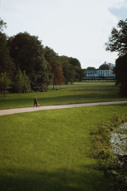 A person walking a small dog along a paved pathway in a large, green park with lush trees and well-maintained grassy areas. In the background, there is a modern, circular building with large windows,
